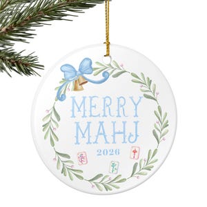 May include: A round white ceramic ornament featuring a green leaf wreath with a light blue bow and gold bells. The text MERRY MAHJ is printed in light blue, with the year 2026 and three small mahjong tile illustrations centered below.