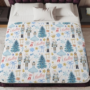 May include: White blanket with a repeating pattern of nutcrackers, ballerinas, Christmas trees, snowflakes, and bows. The name "Hadley" is written in a script font throughout the design. The blanket is on a bed with a purple headboard.