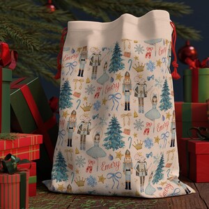 Könnte beinhalten: Ein cremefarbener Weihnachtsgeschenksack mit rotem Kordelzug, der mit einem Muster aus Nussknackern, Weihnachtsbäumen, Schneeflocken und Balletttänzerinnen versehen ist. Der Name "Emery" ist auf der Tasche aufgedruckt. Geschenke und ein Weihnachtsbaum sind im Hintergrund.