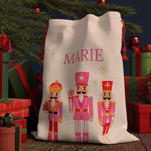 Könnte beinhalten: Ein cremefarbener Weihnachtsgeschenkebeutel mit dem Namen "MARIE" in Rosa und drei Cartoon-Nussknackerfiguren in rosa und roten Uniformen. Der Beutel hat rote Kordelzüge und ist von Geschenken umgeben.