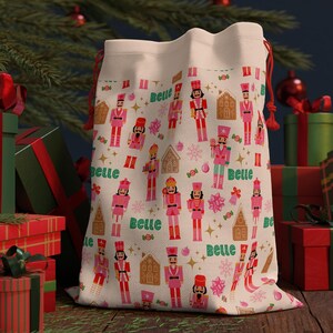 Könnte beinhalten: Ein cremefarbener Weihnachtsgeschenksack mit einem sich wiederholenden Muster aus rosa und roten Nussknackersoldaten, Lebkuchenhäusern und Schneeflocken. Das Wort "Belle" ist ebenfalls auf der Tasche aufgedruckt. Der Beutel ist mit Geschenken gefüllt und hat einen roten Kordelzug.
