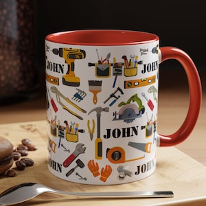Peut inclure: Mug en céramique blanche avec intérieur et anse rouges, orné d'un motif répété d'outils et du nom "JOHN". Les outils comprennent une perceuse, une scie, un marteau et une clé. Le mug est sur une surface en bois avec une cuillère et une pâtisserie.