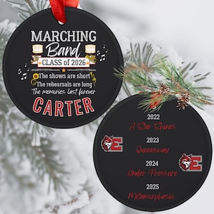 Può includere: Ornamento rotondo nero con nastro rosso. L'ornamento presenta il testo "MARCHING Band CLASS of 2026" e "CARTER". Il retro dell'ornamento mostra gli anni 2022-2025 e il testo "A Star Shines", "Queensway", "Under Pressure" e "Metamorphasis".