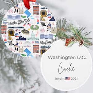 May include: White ceramic ornament with a red ribbon, featuring a Washington D.C. themed design. The design includes illustrations of landmarks, flags, and the text "Washington D.C." and "Cache Intern 2024".