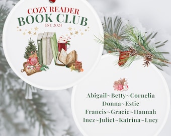 May include: White round ornament with a red ribbon. One side features a watercolor illustration of books, a mug of hot chocolate, and a Christmas tree. The text "Cozy Reader Book Club Est. 2024" is printed in green and red. The other side features a list of names: Abigail, Betty, Cornelia, Donna, Estie, Francis, Gracie, Hannah, Inez, Juliet, Katrina, and Lucy.