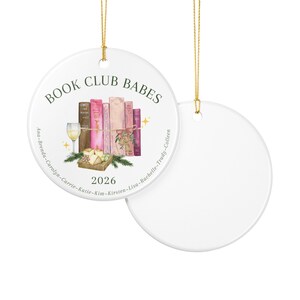 May include: Two round white ceramic ornaments with gold hanging strings. One ornament features the text BOOK CLUB BABES, 2026, and a list of names, with an illustration of books, a wine glass, cheese, and greenery. The other ornament is blank.