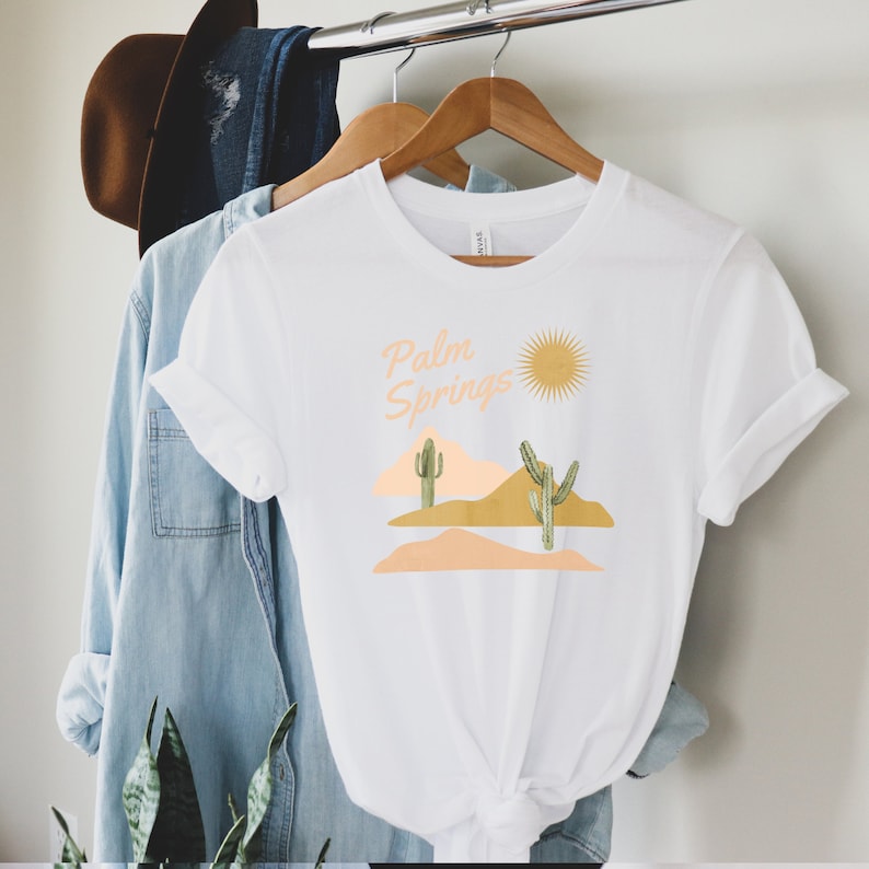 Pu&ograve; includere: Maglietta bianca con un design grafico di una scena del deserto con cactus, montagne e un sole. Il testo "Palm Springs" &egrave; scritto in un font retr&ograve;.