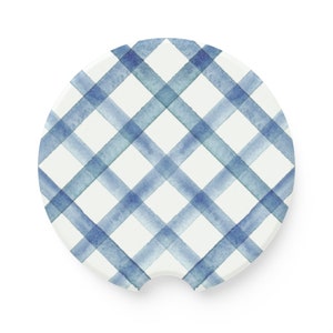 May include: A round car coaster with a white background and a blue plaid pattern. The plaid design is created with watercolor-style brushstrokes in varying shades of blue, creating a soft, textured appearance. The coaster has a small notch on one side.