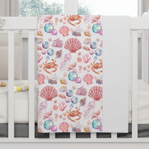 May include: A white baby blanket with a pink, blue, and gold watercolor pattern of seashells, starfish, seahorses, crabs, and bubbles.