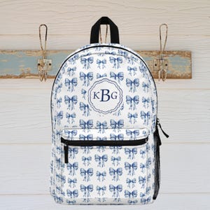 May include: White backpack with a blue bow pattern and a black zipper. The backpack has a black handle and a side mesh pocket. A circular emblem with the initials "KBG" is centered on the front.