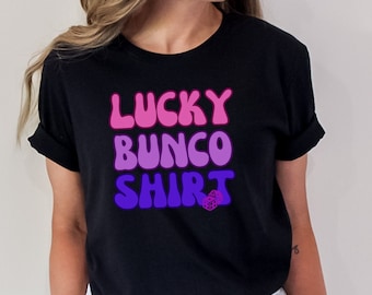 Puede incluir: Una camiseta negra con el texto "LUCKY BUNCO SHIRT" en una fuente retro y colorida. Un pequeño gráfico de dados está debajo del texto.