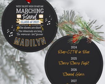May include: Two round black ornaments with red ribbons. The first features West Bluff High Spartan Marching Band Class of 2027, musical notes, instruments, and the name Madilyn. The second lists years 2024-2027 with titles Rhap-CITY in Blue, Starry Starry Night, Stained Glass, and Tango Nuevo.