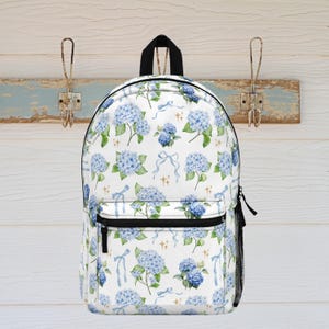 Peut inclure: Sac à dos blanc orné d'un motif floral d'hortensias bleus. Le sac à dos possède une poche zippée noire, une poignée noire et une poche latérale en filet noir. Le motif floral comprend des fleurs bleues, des feuilles vertes et des rubans bleus.