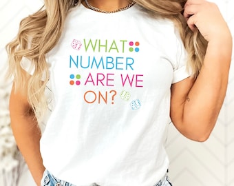 Könnte beinhalten: Weißes T-Shirt mit dem Text "WHAT NUMBER ARE WE ON?" in bunten Buchstaben und Würfelgrafiken.