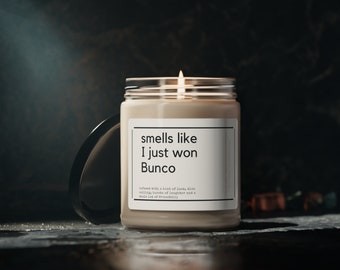 May include: A beige candle with a clear glass jar and a black lid. The candle has a white label with the text "smells like I just won Bunco" and the text "infused with a hint of luck, dice rolling, bursts of laughter and a whole lot of friendship."