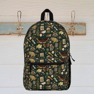 Könnte beinhalten: Ein dunkelgrüner Rucksack mit einem sich wiederholenden Muster aus Schmetterlingen, Fröschen, Pilzen, Briefmarken und Blumen. Der Rucksack hat einen schwarzen Reißverschluss, einen schwarzen Griff und eine Seitentasche aus Netz. Er hängt an einer rustikalen Wand.