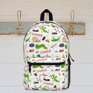 May include: A white backpack featuring a repeating pattern of various colorful insects, snails, and worms, interspersed with the name Charlie printed in red text. The bag includes a black top handle, black zippers, and a black mesh side pocket.
