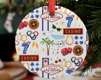 May include: A round, white Christmas ornament with a Las Vegas theme. The ornament features a repeating pattern of casino-related icons, including dice, playing cards, and the words "Welcome to Fabulous Las Vegas Nevada". A red ribbon is attached for hanging.