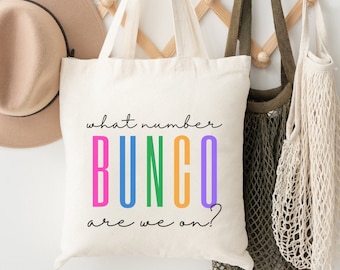 May include: A white canvas tote bag with the text "what number BUNCO are we on?" in black and rainbow colors. The word BUNCO is in large letters with each letter in a different color: pink, blue, green, orange, and purple.