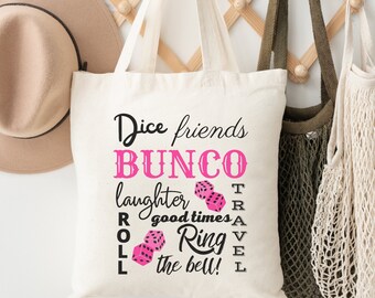Op de afbeelding: Een witte canvas tote bag met zwarte tekst die "Dice friends BUNCO laughter good times ROLL Ring the bell! TRAVEL" luidt. De tekst wordt omringd door roze dobbelstenen.