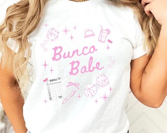 Op de afbeelding: Wit T-shirt met een roze grafisch ontwerp met de tekst "Bunco Babe". Het ontwerp bevat dobbelstenen, sterren, een cocktailglas, een zon en een scorekaart.