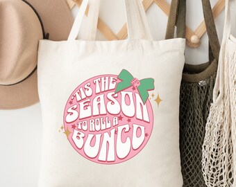 Op de afbeelding: Een witte canvas tote bag met een roze cirkelvormig ontwerp met de tekst "Tis the season to roll a Bungo". Het ontwerp bevat ook een groene strik met een dobbelsteen en gouden sterren.