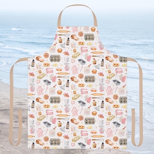 May include: A white apron featuring a repeating pattern of baking-themed illustrations in pink, beige, and brown. The pattern includes rolling pins, cupcakes, and baking pans. The apron has a beige neck strap and waist ties.