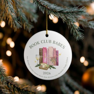 May include: A round white ceramic ornament featuring the text BOOK CLUB BABES at the top and 2026 at the bottom. The center displays a watercolor illustration of five books, a wine glass, and a cheese board. Names are printed along the edge.