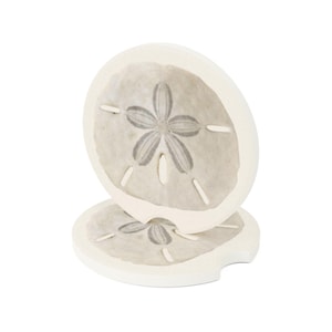 May include: Two round white coasters featuring a gray sand dollar design with a central five-petal star pattern and small elongated slits. The coasters have a notched edge and a smooth, flat surface, presented with one standing upright.