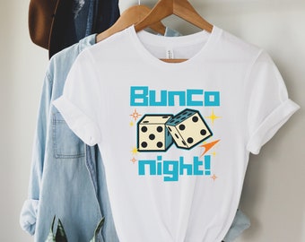 Puede incluir: Camiseta blanca con el texto "Bunco night!" en turquesa, con dos dados. Los dados son de color blanco roto con puntos negros y un diseño retro. La camiseta está colgada en una percha de madera.