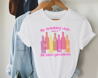Puede incluir: Camiseta blanca con un gráfico rosa que dice "My drinking club has a BUNGO problem". El gráfico presenta una fila de botellas de vino coloridas con dados sobre ellas.