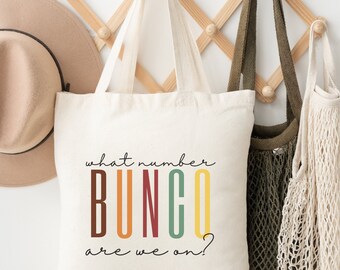 Op de afbeelding: Een witte canvas draagtas met de tekst "what number BUNCO are we on?" in zwart gedrukt. Het woord "BUNCO" staat in kleurrijke blokletters met rood, oranje, geel, groen en blauw.