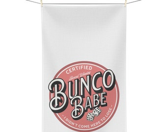 Peut inclure: Serviette de cuisine blanche gaufrée avec un motif circulaire rouge et blanc. Le motif indique "BUNCO BABE" avec des dés et le texte "I DIDN'T COME HERE TO LOSE". La serviette est suspendue à une corde à linge.