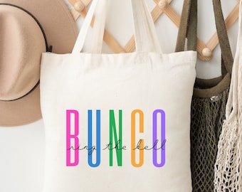 Op de afbeelding: Een witte canvas tote bag met het woord "BUNCO" bedrukt in regenboogkleuren. De tekst "ring the bell" is in zwarte cursieve letters onder het woord "BUNCO" geschreven.