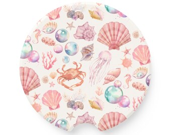 Può includere: Un sottobicchiere bianco con un design ad acquerello di conchiglie, stelle marine, meduse e bolle. Il design è in tonalità di rosa, blu e verde.