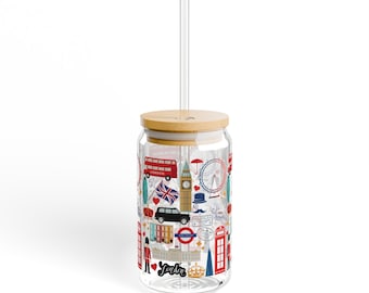 May include: A clear glass can with a bamboo lid and a straw. The can is decorated with a London theme, featuring images of a red double-decker bus, Big Ben, the London Eye, and other iconic landmarks. The can says "London" on the bottom.