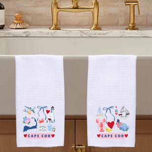 Puede incluir: Dos paños de cocina blancos con tejido de gofre con diseños coloridos con temática de Cape Cod. Cada paño presenta un faro, una langosta, un velero y el texto "CAPE COD" con corazones. Los paños cuelgan frente a un fregadero.