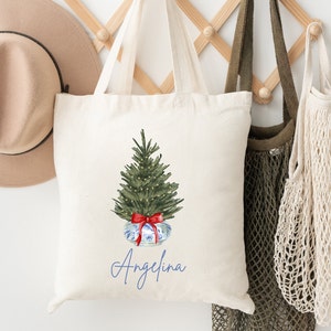May include: A white canvas tote bag with a green Christmas tree with white lights and a red bow. The tree is in a blue and white patterned pot. The name "Angelina" is written in blue cursive below the tree.