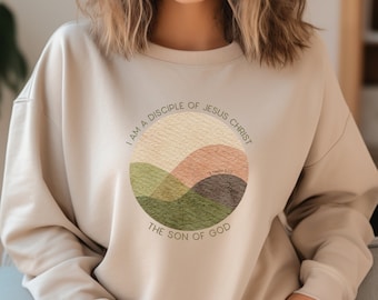 May include: Beige sweatshirt with a round graphic design featuring a stylized landscape in shades of green, brown, and beige. The text "I am a disciple of Jesus Christ" is written in a circle around the graphic, with the text "The Son of God" below the graphic. The text "1 Timothy 6:13" is written in small font within the graphic.