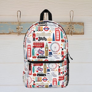 May include: White backpack with a repeating pattern of London landmarks. The design includes red buses, telephone boxes, the London Eye, Big Ben, and the word "London". The backpack has a black handle and a front pocket.