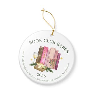 May include: A round white ceramic ornament with a gold hanging string. The design features a stack of pink and brown books, a wine glass, and cheese. Text reads BOOK CLUB BABES, 2026, and a list of names: Ana-Brenda-Carolyn-Carrie-Katie-Kim-Kirsten-Lisa-Rachelle-Trudy-Colleen.