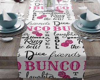 Peut inclure: Un chemin de table blanc avec un motif rose et noir représentant les mots "Bunco", "Dés", "Amis", "Rire", "Bons moments", "Sonner la cloche" et "Voyage". Le motif comprend également des dés avec des points roses.