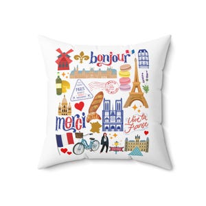 May include: A white pillow with a colorful design featuring Parisian landmarks, including the Eiffel Tower, Notre Dame Cathedral, and a baguette. The pillow also includes the words "bonjour", "merci", and "vive la France".