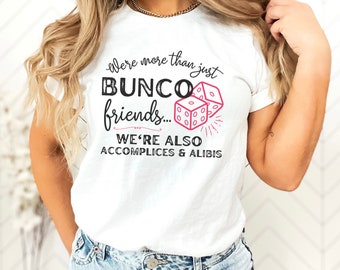 Puede incluir: Camiseta blanca con el texto "We're more than just Bunco friends... We're also accomplices & alibis." El diseño incluye dados rosas y una explosión decorativa.