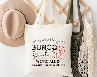 Op de afbeelding: Witte canvas tote bag met zwarte tekst die "We're more than just Bunco friends... We're also accomplices & alibis" luidt. De tekst is in een vintage lettertype. Er zijn rode dobbelstenen met witte stippen op de tas.