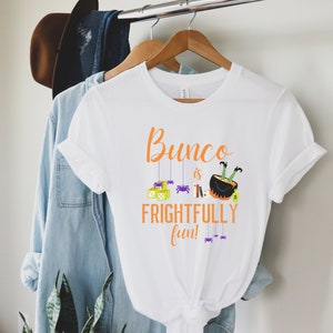 May include: White t-shirt with an orange graphic that says "Bunco is Frightfully Fun!"  The graphic includes a witch's cauldron with legs, dice, and spiders.