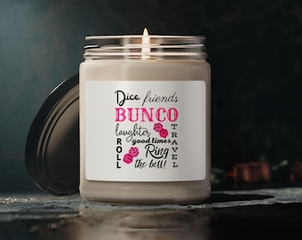 Może przedstawiać: Biała świeca z brązową etykietą, na której widnieje napis "Dice friends BUNCO laughter good times ROLL Ring the bell!" z różowymi kostkami do gry i słowami "TRAVEL" w kolorze czarnym.