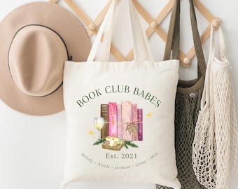 Puede incluir: Un bolso de tela color crema con el texto "BOOK CLUB BABES" en verde oscuro, una ilustración de cinco libros en tonos rosa y marrón, una copa de vino blanco, una pequeña tabla de quesos y el texto "Est. 2021" con cinco nombres debajo.