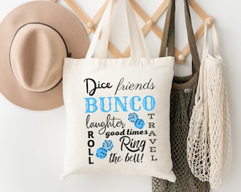 Op de afbeelding: Een crèmekleurige draagtas met de tekst "Dice friends BUNCO laughter good times ROLL Ring the bell!" in zwart en blauw. De tas heeft dobbelsteenillustraties. Een beige hoed en twee andere tassen hangen aan een houten rek.