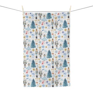 May include: A white hand towel with a repeating pattern of blue Christmas trees, nutcrackers, ballerinas, bows, and snowflakes. The towel is hanging from a clothesline with a wooden clothespin.